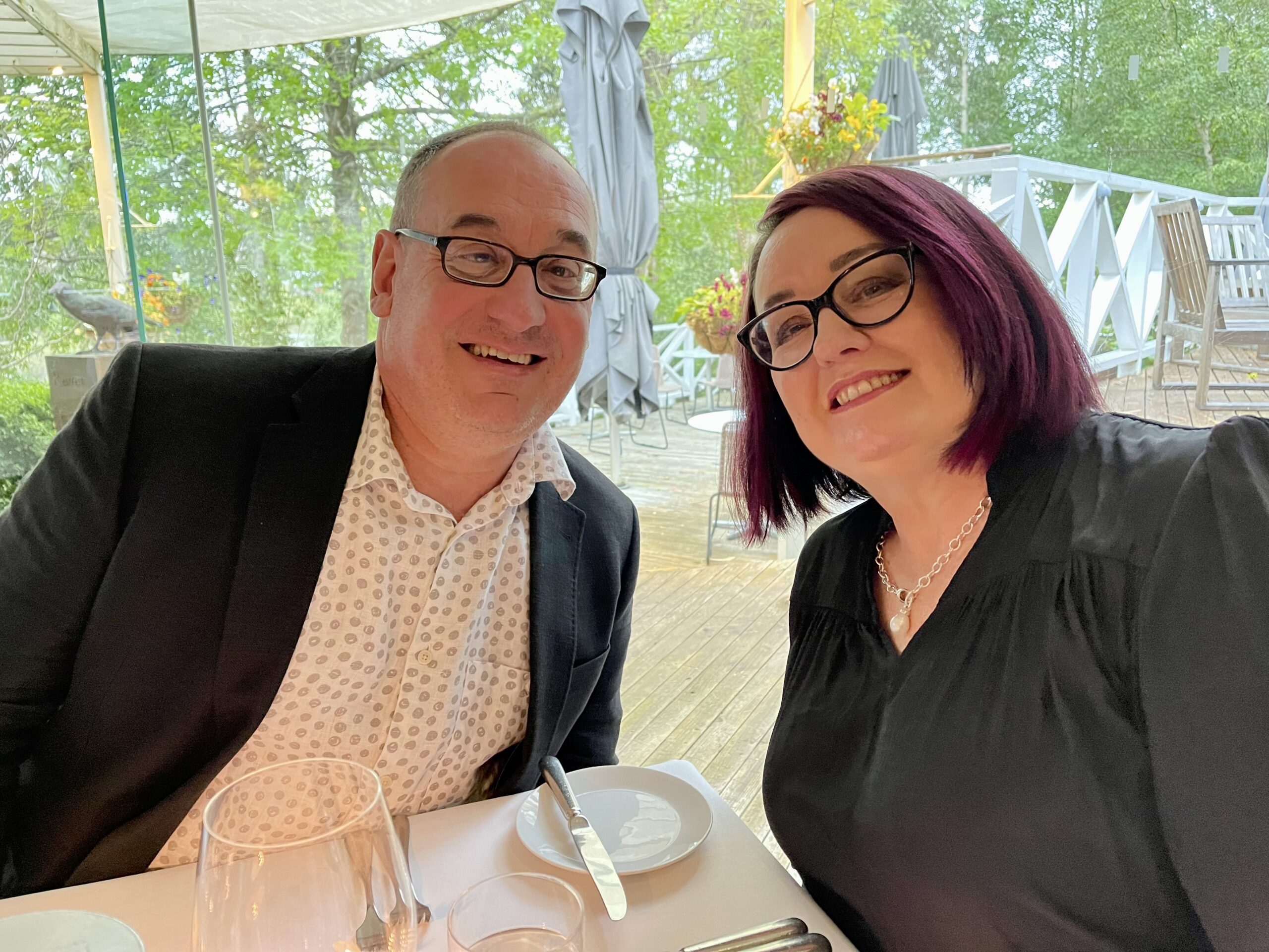 An image of Michael, left and Mardi, right. Michael has a collared shirt, white, with a dark jacket. Mardi is in a black dress wearing jewellery. They both wear glasses and are sitting at a table with white linen.  